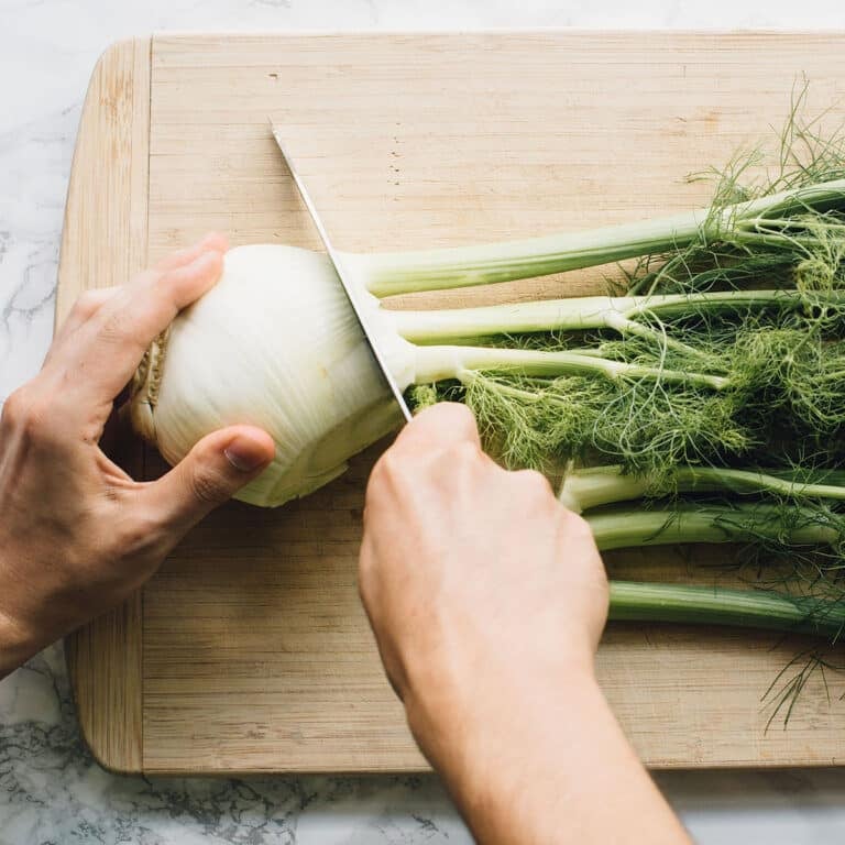 A fennel bulb on a cutting board. The stalks and fronds are being cut away from the bulb.