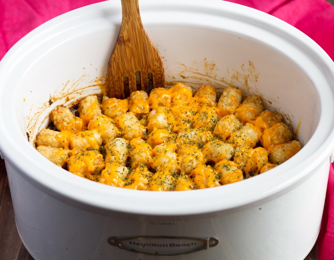 white slow cooker liner filled with Tater Tot Casserole covered in melted yellow cheese and garnished with finely chopped parsley; there is a wooden spoon sticking out of the slow cooker.