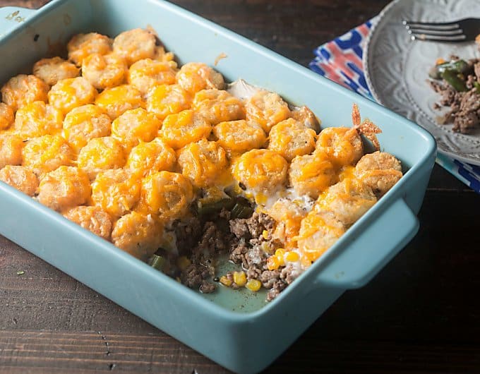 light blue ceramic casserole dish filled with Tater Tot Casserole topped with melted cheddar cheese; a scoop is missing from the casserole.