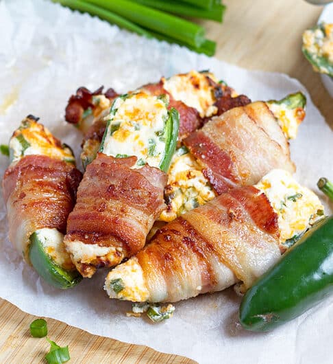 jalapenos stuffed with white filling with specks of green and orange cheddar, wrapped in bacon on parchment paper and small white plate; beige cloth with black dots in background