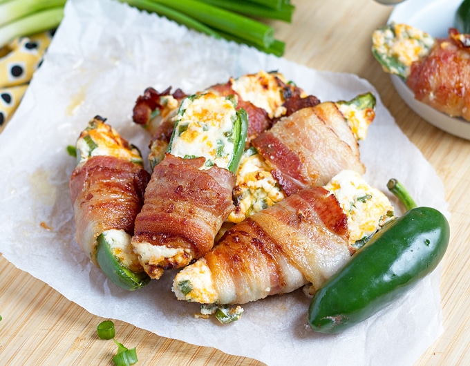 jalapenos stuffed with white filling with specks of green and orange cheddar, wrapped in bacon on parchment paper and small white plate; beige cloth with black dots in background