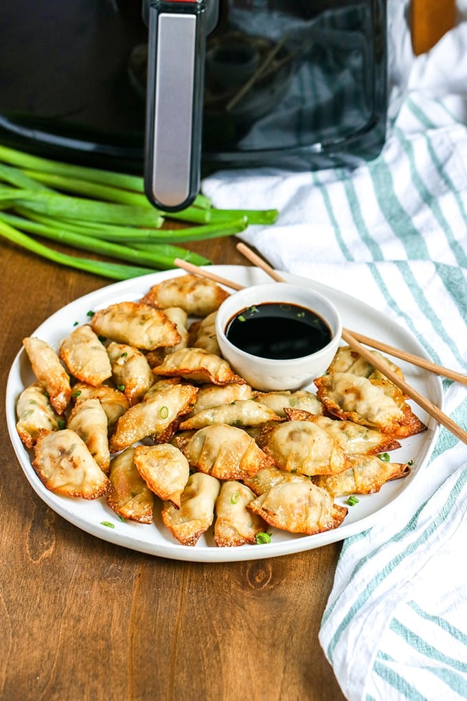 white plate full of pot stickers with condiment bowl of soy sauce and chopsticks; white cloth with blue stripes to right side with air fryer and green onion in background