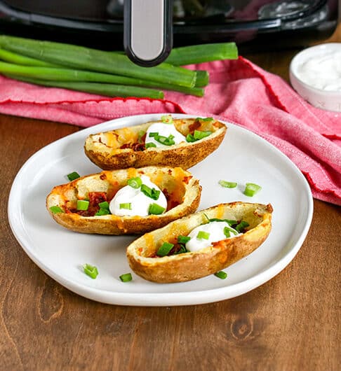 potato skins on white plate with green onion and sour cream, pink cloth behind plate with green onions, sour cream and air fryer in background