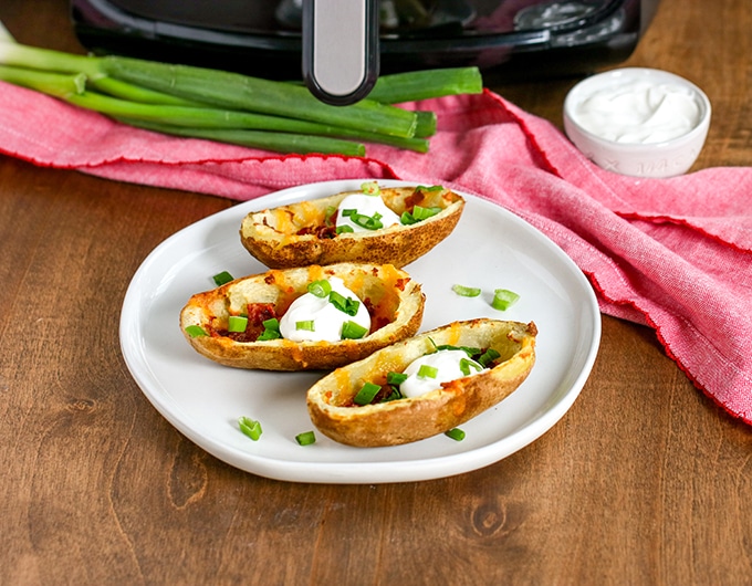 potato skins on white plate with green onion and sour cream, pink cloth behind plate with green onions, sour cream and air fryer in background