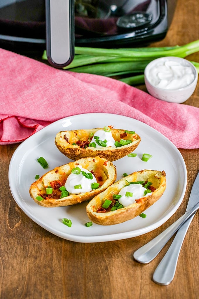 potato skins on white plate with green onion and sour cream; silverware off to right handside, pink cloth behind plate with green onions, sour cream and air fryer in background