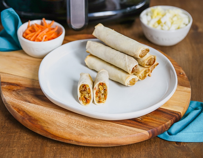 stack of 5 spring rolls on white plate on top of wooden platter; 1 spring roll cut in half on front half of plate; small bowl of shredded carrots, back left with air fryer behind it; small bowl with cabbage shreds in white bowl to back right