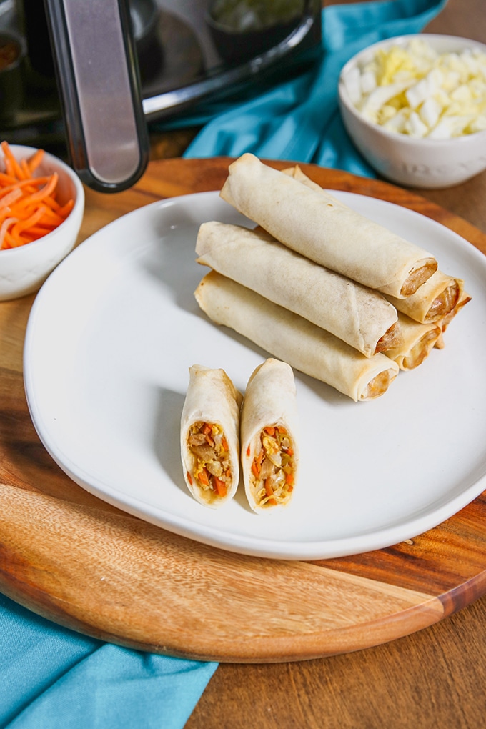 stack of 5 spring rolls on white plate on top of wooden platter; 1 spring roll cut in half on front half of plate; small bowl of shredded carrots, back left with air fryer behind it; small bowl with cabbage shreds in white bowl to back right