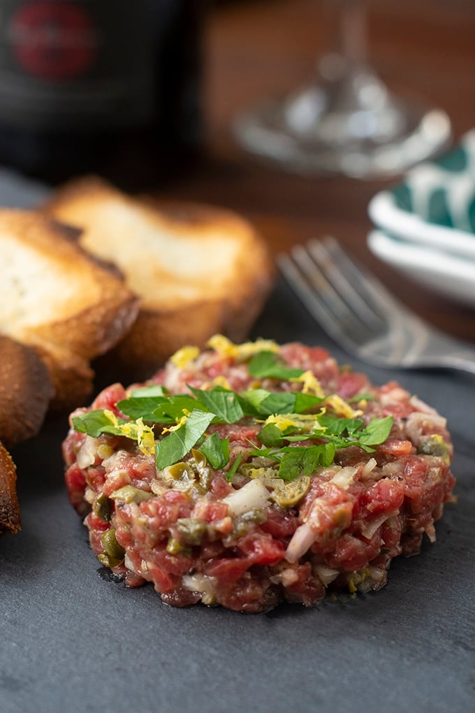 Raw beef finely chopped mixed with finely chopped onions and capers, shaped into a patty and topped with chopped basil and lemon zest. In the background you can see some toasted bread and a fork.