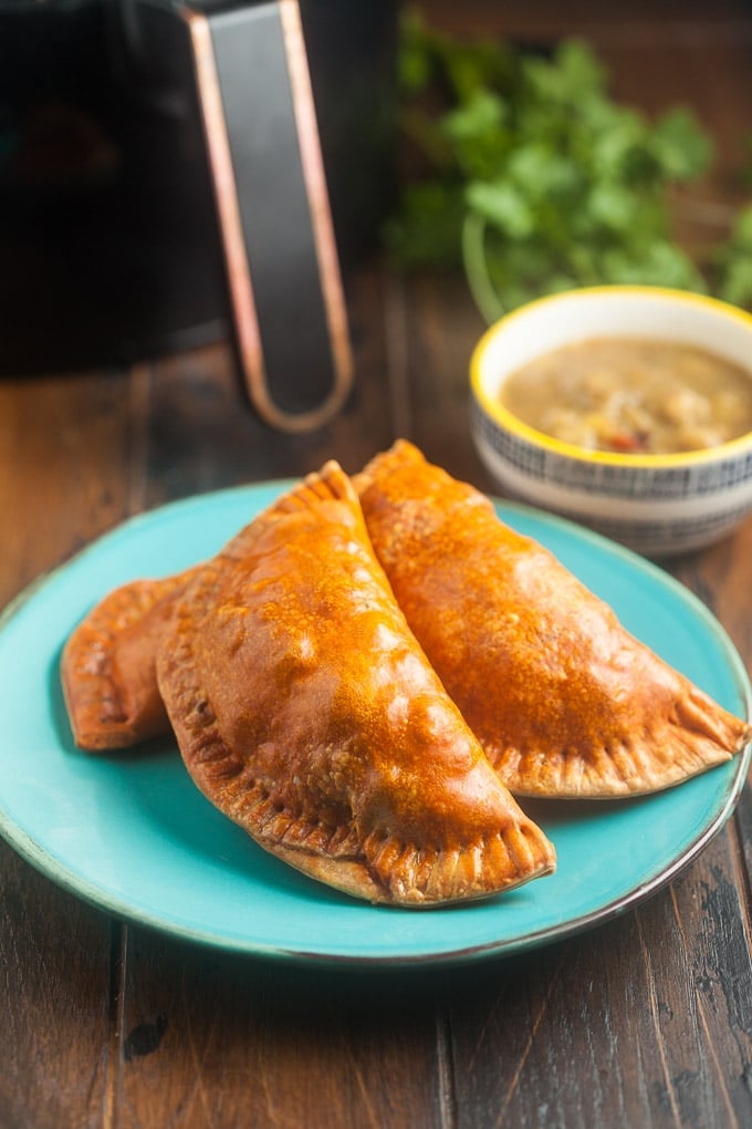 3 empanadas on a bright blue plate sitting on a wood table in front of an air fryer and a bowl filled with a light brown stew.