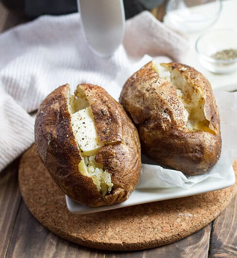 2 baked potatoes cut open with butter and pepper on white tile on a piece of cork; white cloth and air fryer in background