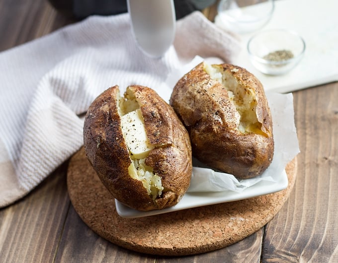 2 baked potatoes cut open with butter and pepper on white tile on a piece of cork; white cloth and air fryer in background