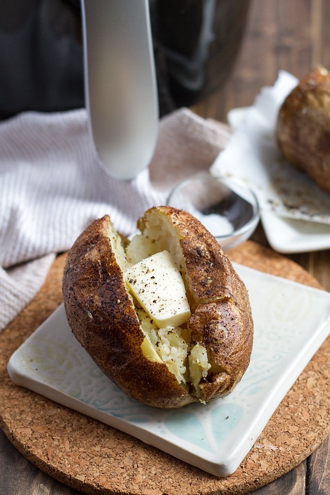 baked potato cut open with butter and pepper on white and light blue tile on a piece of cork; white cloth and air fryer in background