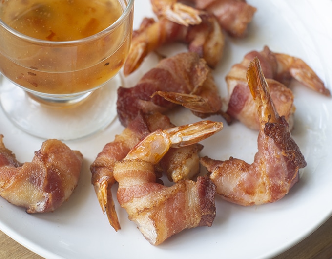 A bunch of crispy bacon wrapped shrimp on a round white plate with a glass cup of golden dipping sauce.