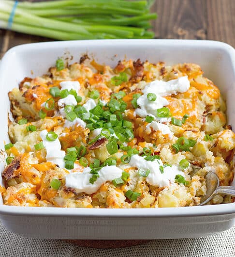 white casserole dish with potato topped with melted orange cheddar, sour cream, green onion, and bacon bits; beige cloth underneath and green onion the background; metal spoon in dish