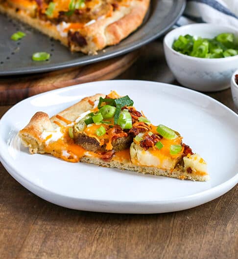 Loaded baked potato pizza in background on a metal pizza tray, one slice missing and is plated in front on white plate topped with sour cream, melted orange cheddar, green onion, and bacon bits; behind in white condiment bowls garnish of green onion and bacon bits