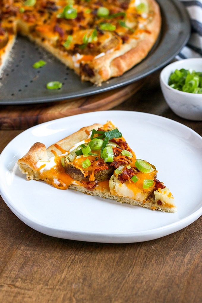 Loaded baked potato pizza in background on a metal pizza tray, one slice missing and is plated in front on white plate topped with sour cream, melted orange cheddar, green onion, and bacon bits; behind in white condiment bowl garnish of green onion