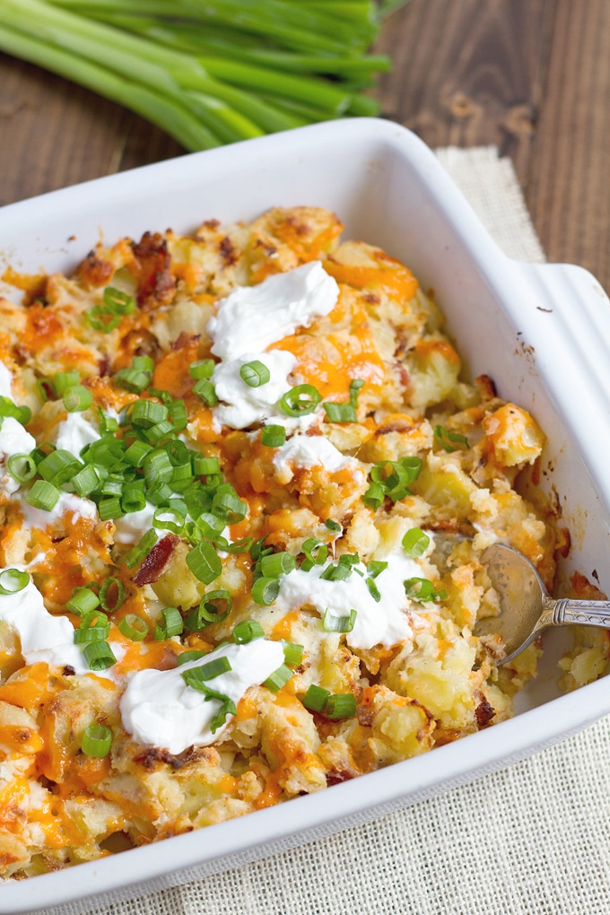 white casserole dish with potato topped with melted orange cheddar, sour cream, green onion, and bacon bits; beige cloth underneath and green onion the background