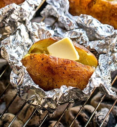 3 baked potatoes in foil; one near front of image has butter; all are on grill grate over white coals