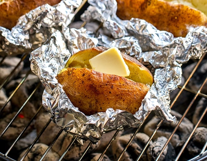 3 baked potatoes in foil; one near front of image has butter; all are on grill grate over white coals