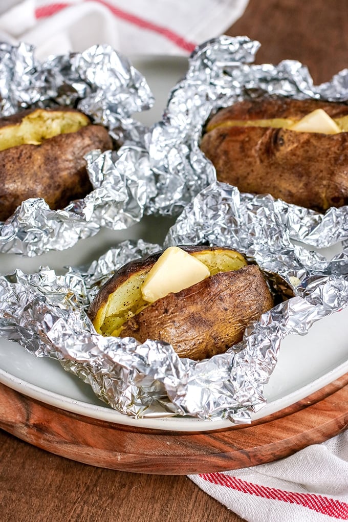 3 baked potatoes in foil on white platter on top of wooden tray; white cloth with red stripe under tray; potato closest to front of image is cut open with butter and pepper