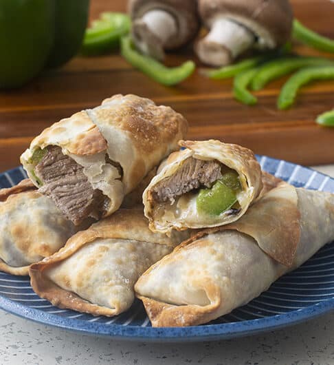steak, cheese, and bell pepper showing from cut air fried egg roll on stack of egg rolls on medium colored blue plate; green bell pepper whole and sliced and 2 mushrooms on cutting board in background