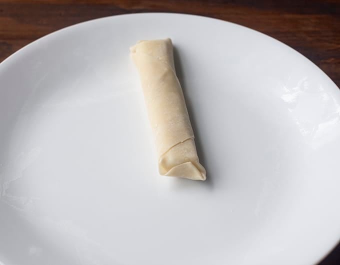 egg roll wrapper wrapped around cheese stick on white plate showing end folded down and pinched closed
