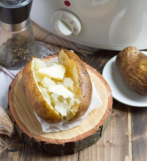 baked potato with butter on foil on wooden log round with beige and black plaid cloth underneath; another baked potato uncut on small white plate; salt and pepper and white slow cooker in background