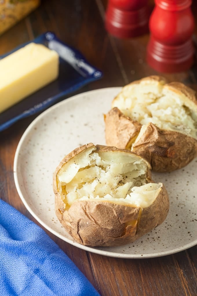 2 baked potatoes cut open on light tan plate with black speckles, salt and pepper; top right bottom of red salt shaker showing; top left butter on dark blue dish; bottom left blue cloth