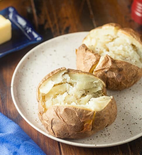 2 baked potatoes cut open on light tan plate with black speckles, salt and pepper; top right bottom of red salt shaker showing; top left butter on dark blue dish; bottom left blue cloth