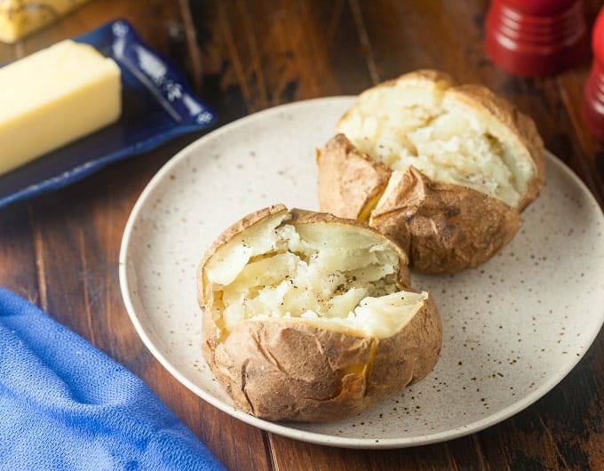 2 baked potatoes cut open on light tan plate with black speckles, salt and pepper; top right bottom of red salt shaker showing; top left butter on dark blue dish; bottom left blue cloth