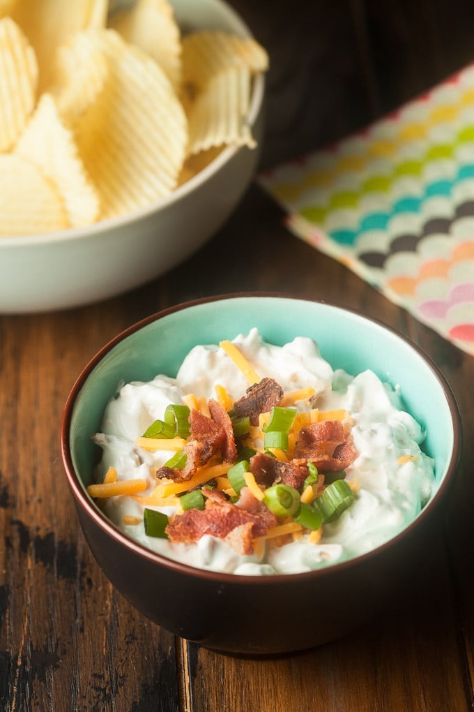 Loaded baked potato dip with orange cheddar, green onion, and bacon bits topping it in bowl that is brown outside and light blue inside; white bowl with rippled potato chips in top left corner; right corner has cloth with wave stripe in yellow, green, blue, black, orange, pink