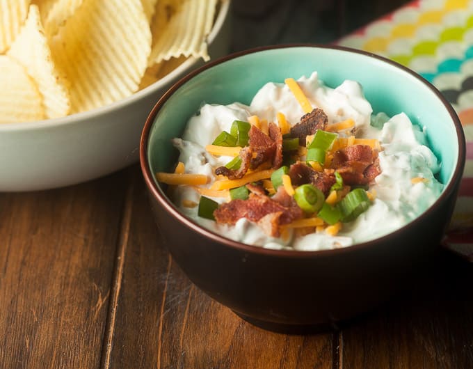 Loaded baked potato dip with orange cheddar, green onion, and bacon bits topping it in bowl that is brown outside and light blue inside; white bowl with rippled potato chips in top left corner; right corner has cloth with wave stripe in yellow, green, blue, black, orange, pink