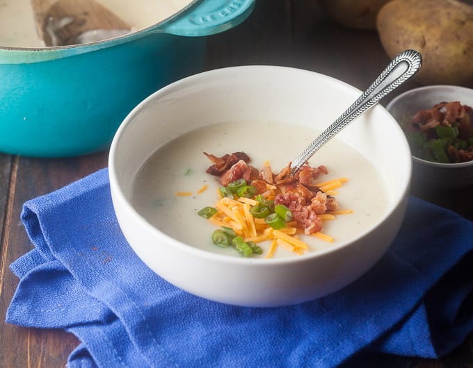 potato soup in white bowl on blue cloth with green onion, orange cheddar, bacon bits, and spoon in bowl; small bowl behind soup with bacon bits; left hand top corner has light blue dutch oven