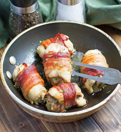 5 chicken thighs wrapped in bacon with parsley garnish in skillet; sage colored cloth in background and salt and pepper grinder in background on dark brown wooden table; meat fork in pan