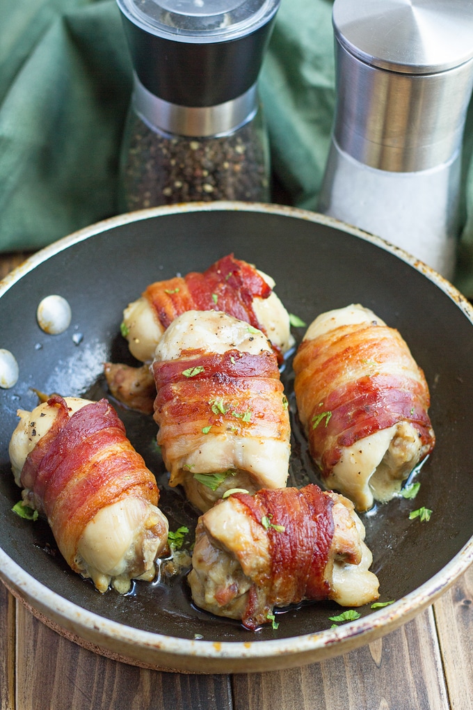 5 chicken thighs wrapped in bacon with parsley garnish in skillet; sage colored cloth in background and salt and pepper grinder in background on dark brown wooden table