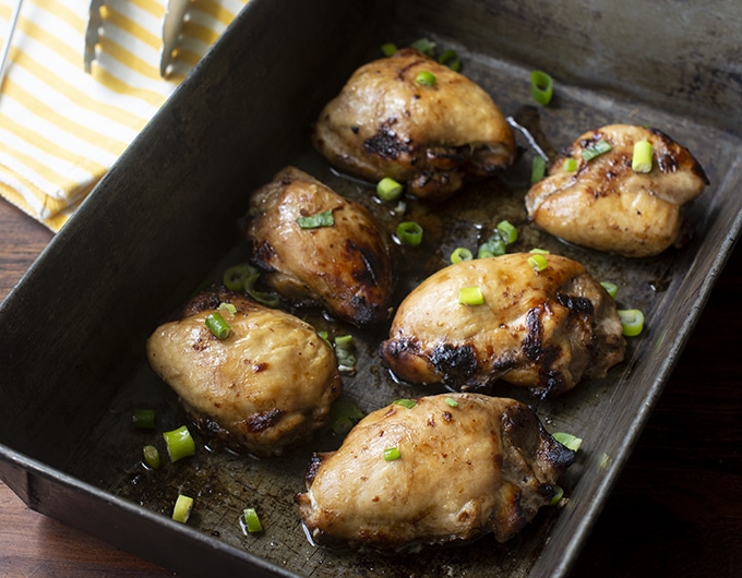 baked chicken thighs in metal baking pan with green onion garnish; yellow and white striped towel and tongs in background