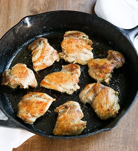 browned chicken thighs in large lodge cast iron skillet; white cloth with red stripe underneath skillet; on brown wood table
