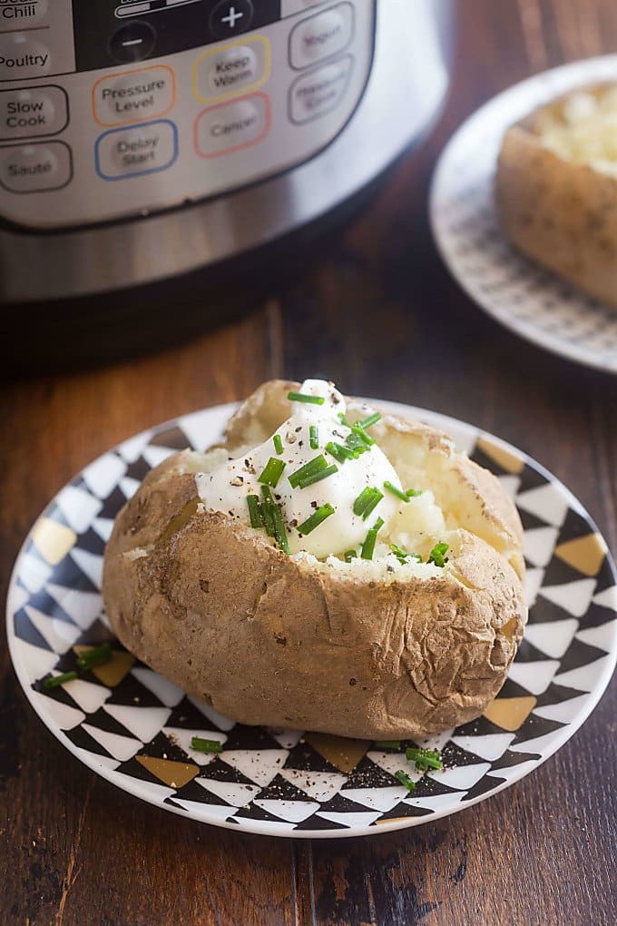 2 baked potatoes with sour cream and chives on white plates; one plate with black and gold triangles and the other with black and gold dots; bottom of Instant Pot in background