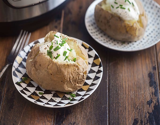 2 baked potatoes with sour cream and chives on white plates; one plate with black and gold triangles and the other with black and gold dots; fork to left of plate and small corner of Instant Pot in background