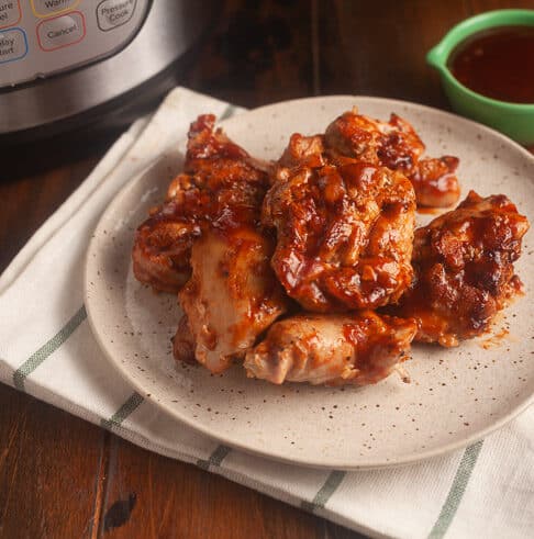 white plate with black speckles on white cloth with black stripes; chicken thighs with bbq sauce on plate; instant pot in background and light green condiment bowl with bbq sauce in it in background