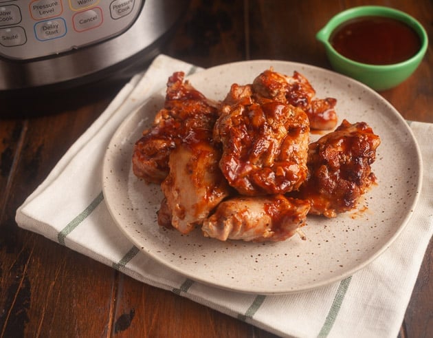white plate with black speckles on white cloth with black stripes; chicken thighs with bbq sauce on plate; instant pot in background and light green condiment bowl with bbq sauce in it in background