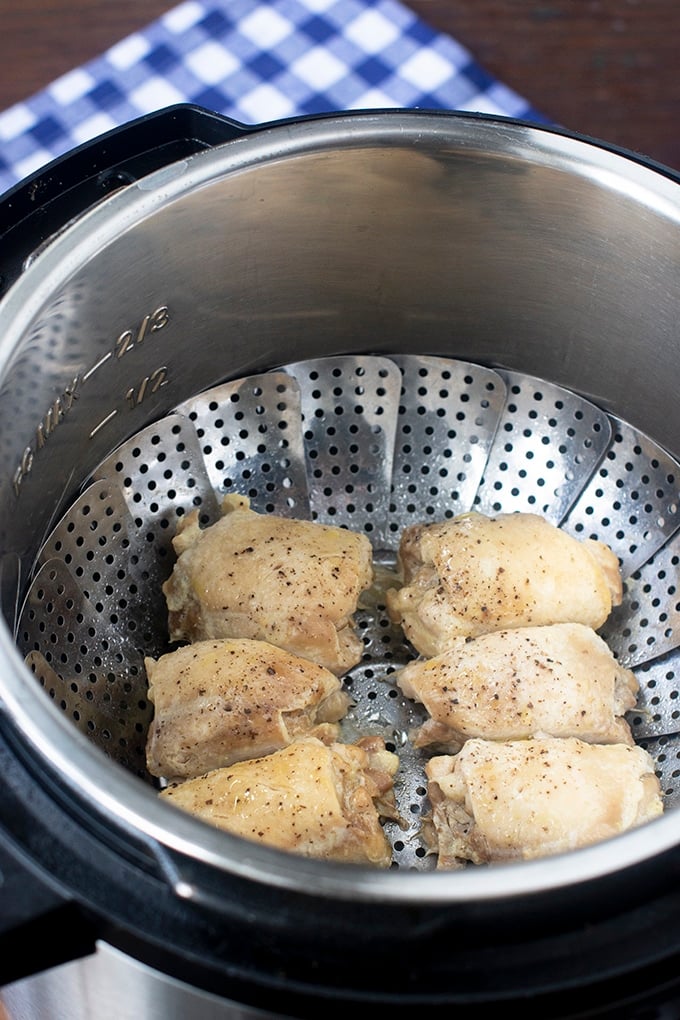 6 chicken thighs with black pepper on them in stainless steel steamer basket in instant pot insert; blue and white checked cloth in background
