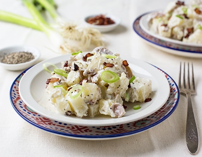 potato salad with black pepper, green onion, and bacon bits on white plate on red, white, and blue charger; fork to right of plate and small white condiment bowl with what looks like black pepper to back lect, green onions in background and part of another plate of potato salad in background corner
