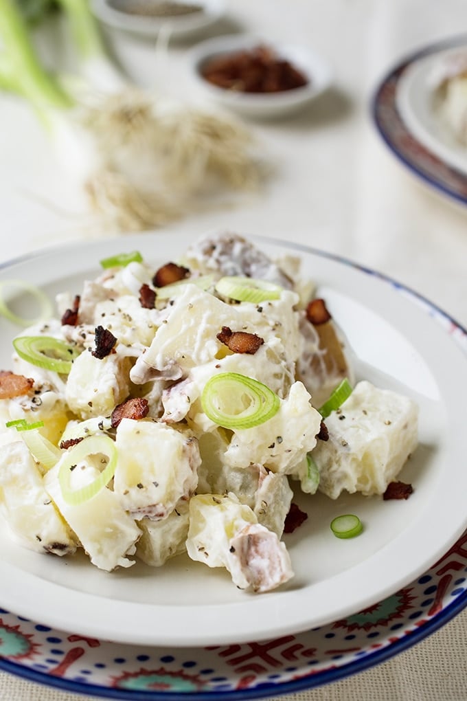 potato salad with black pepper, green onion, and bacon bits on white plate on red, white, and blue charger; small white condiment bowl with bacon bits behind plate , green onions in background and part of another plate of potato salad in background corner