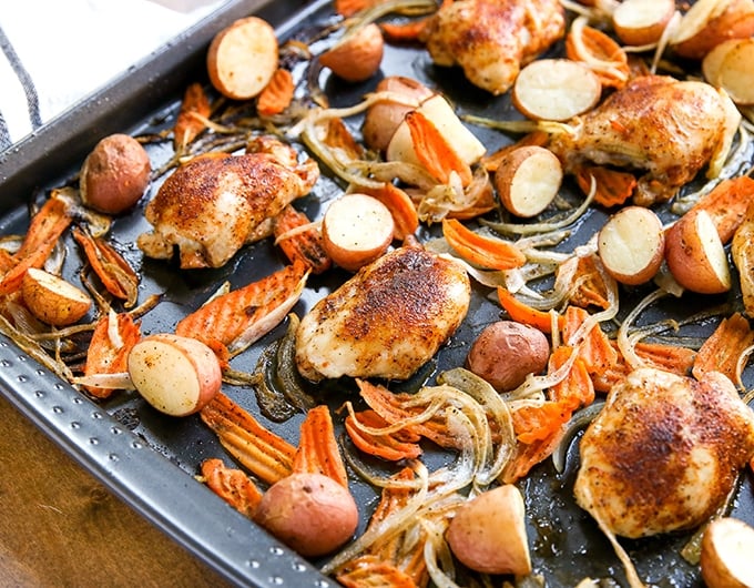 sheet pan with halved baby red potatoes, carrot chips, onion slices, and seasoned chicken thighs