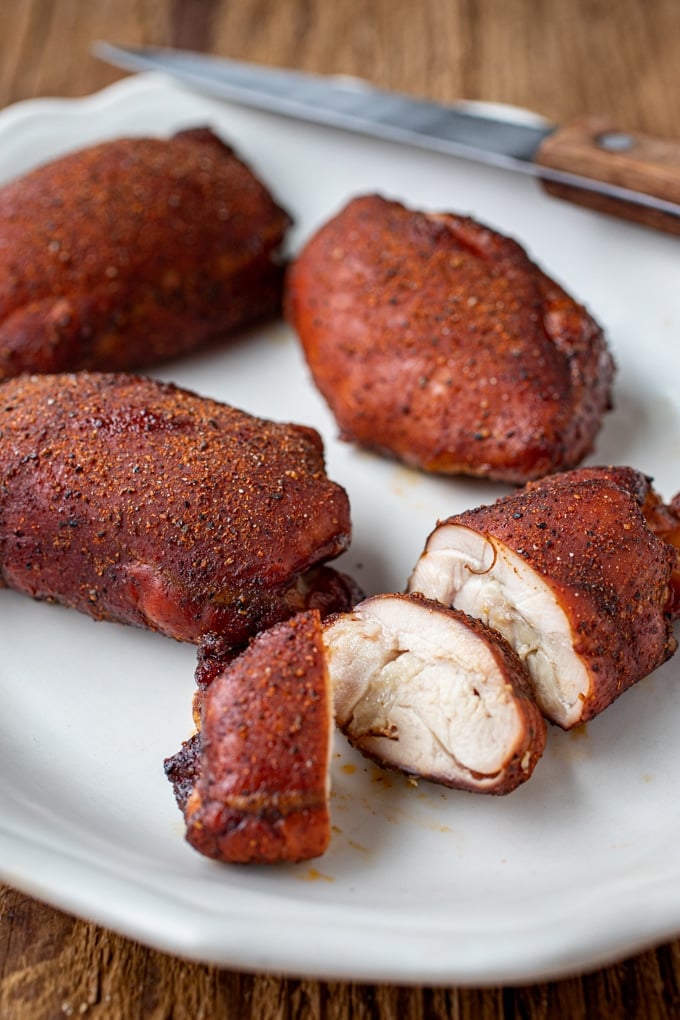 4 smoked chicken thighs coated in seasoning and a burnished red brown; one sliced; on white platter with knife in background on brown wood table