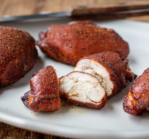 4 smoked chicken thighs coated in seasoning and a burnished red brown; one sliced; on white platter with knife in background on brown wood table