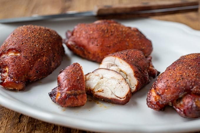 4 smoked chicken thighs coated in seasoning and a burnished red brown; one sliced; on white platter with knife in background on brown wood table