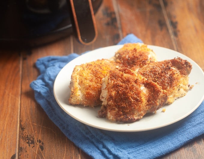 4 chicken thighs breaded with panko on white plate sitting on medium blue colored cloth; bottom of air fryer and handle in background