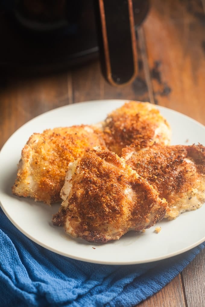 4 chicken thighs breaded with panko on white plate sitting on medium blue colored cloth; bottom of air fryer and handle in background